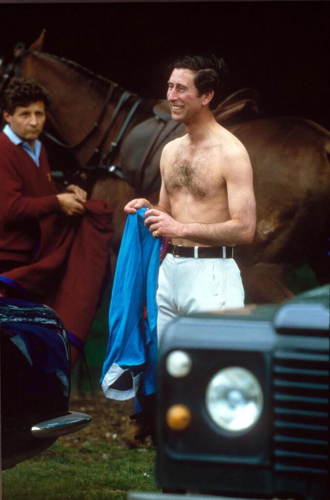 Prince Charles shirtless, smiling, wearing white trousers and holding a blue polo top