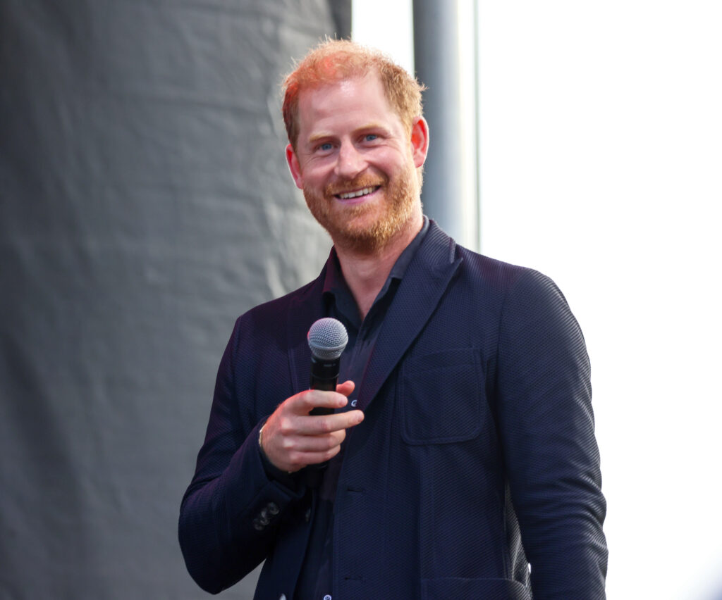 Prince Harry wears a black suit and shirt and stands on a stage