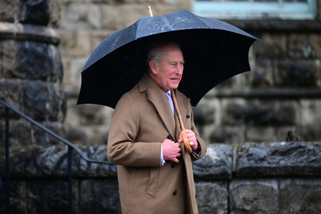 King Charles holding an umbrella whilst wearing a camel coat