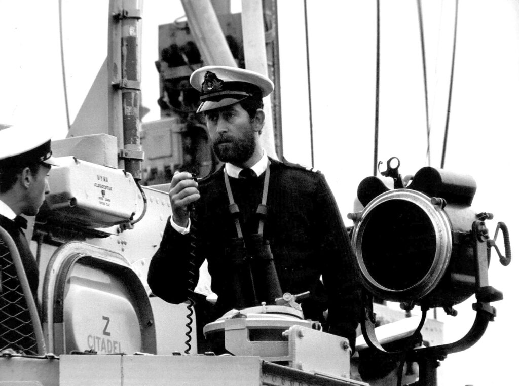 Prince Charles photographed speaking into a radio, wearing a hat and sporting a beard upon a Royal Navy ship
