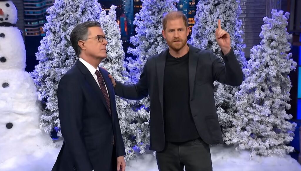 Stephen Colbert and Prince Harry on stage in front of a set of Christmas trees and snow