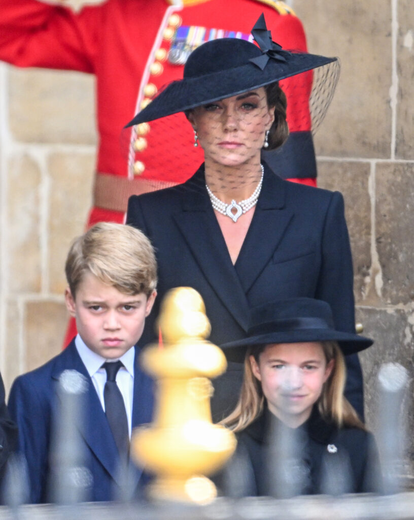 Princess Kate wearing a black hat and coat dress with. pearl choker at The State Funeral of Queen Elizabeth II at Westminster Abbey with Prince George and Princess Charlotte