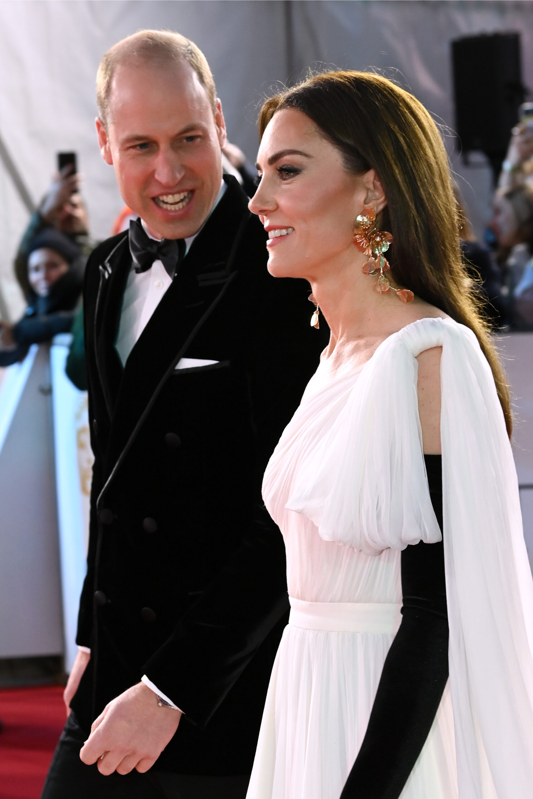 Prince William wearing a tuxedo whilst leaning in and talking to Princess Kate wearing a long white dress, whilst smiling, as they walk on the red carpet, at the BAFTAs in 2023