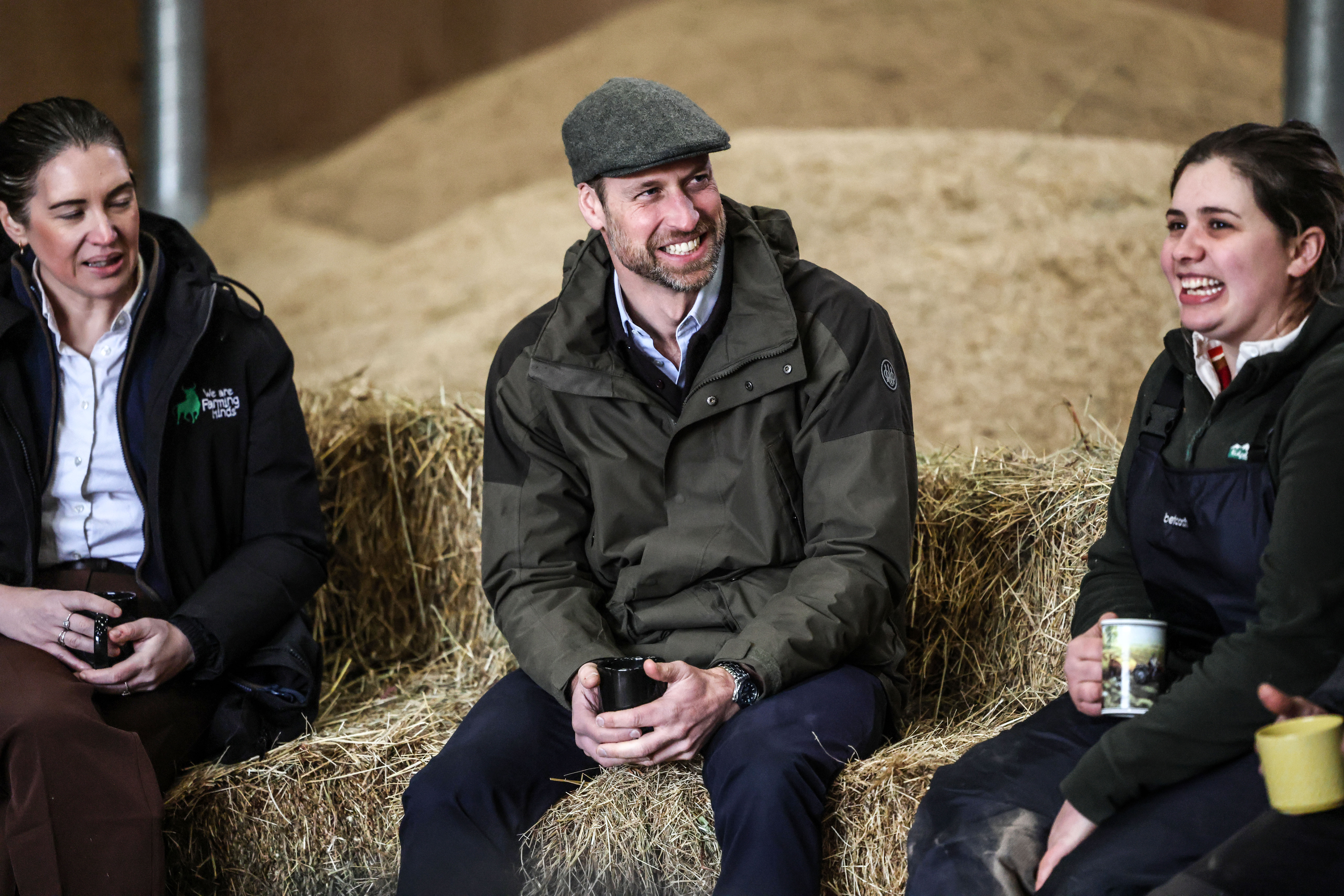 Prince William smiling wearing a flat cap and a green coat sat on a hay bale
