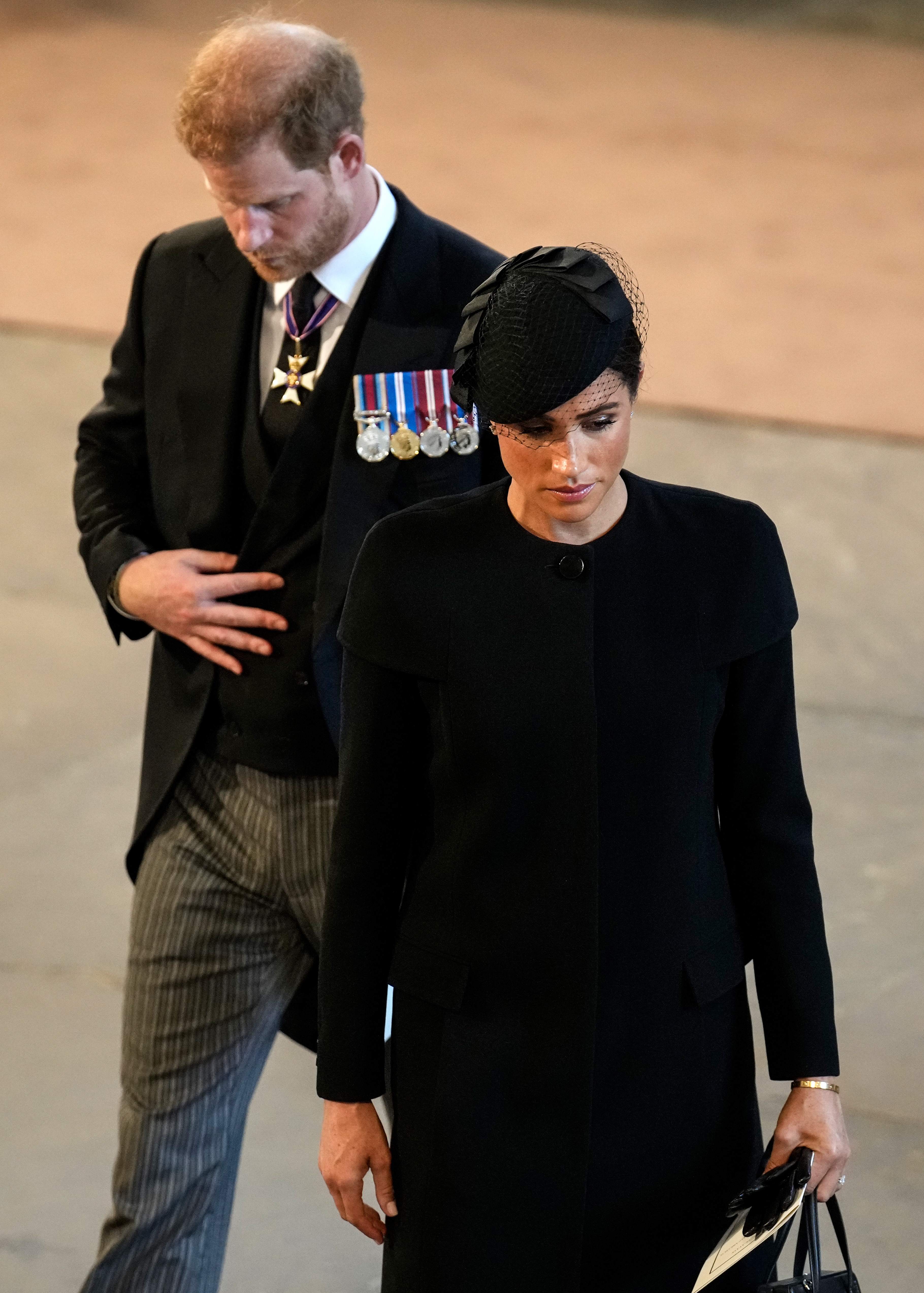 Prince Harry looking down as he walks behind Meghan Markle frowning