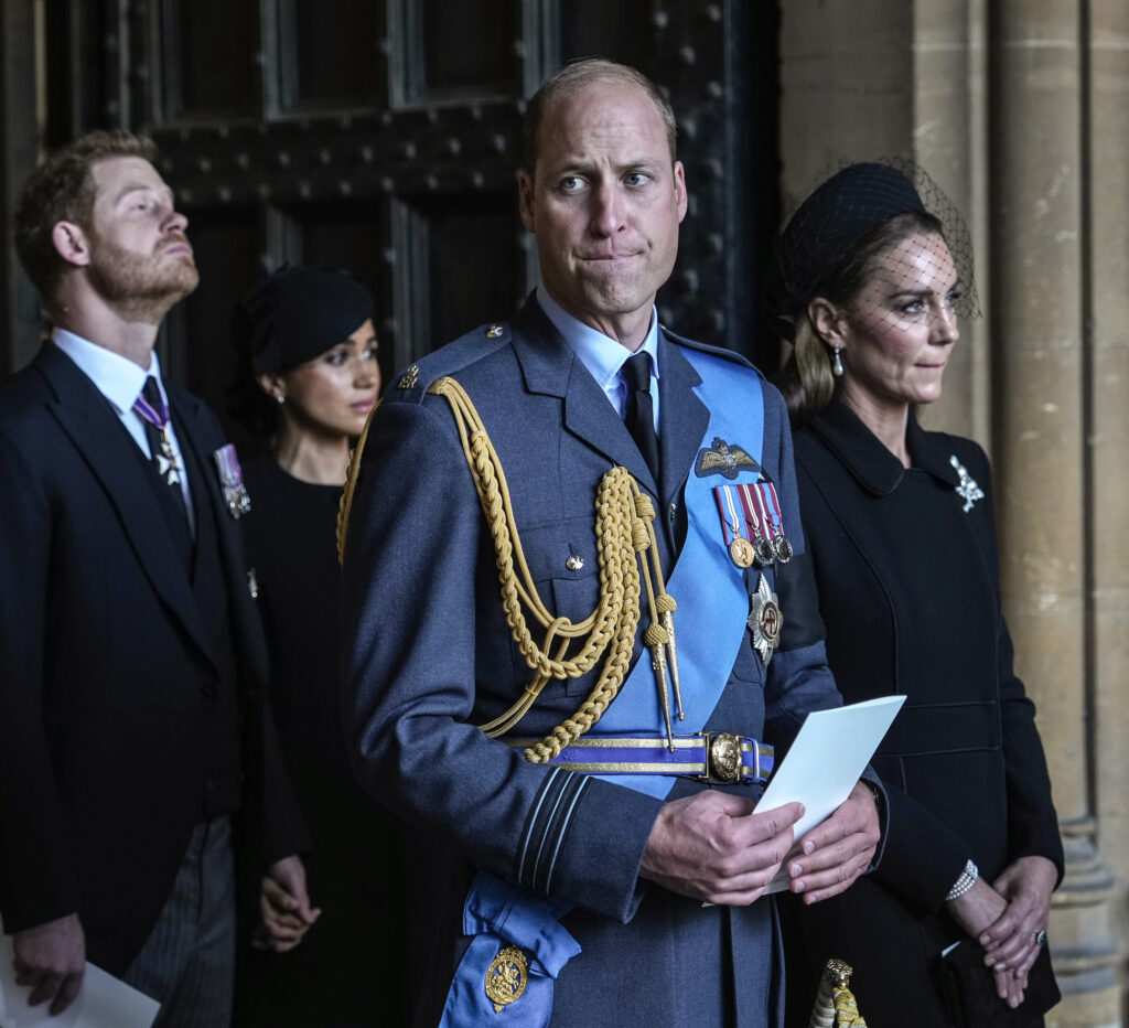 Prince Harry beside Meghan Markle, both frowning, behind Prince William and Princess Kate with serious expressions