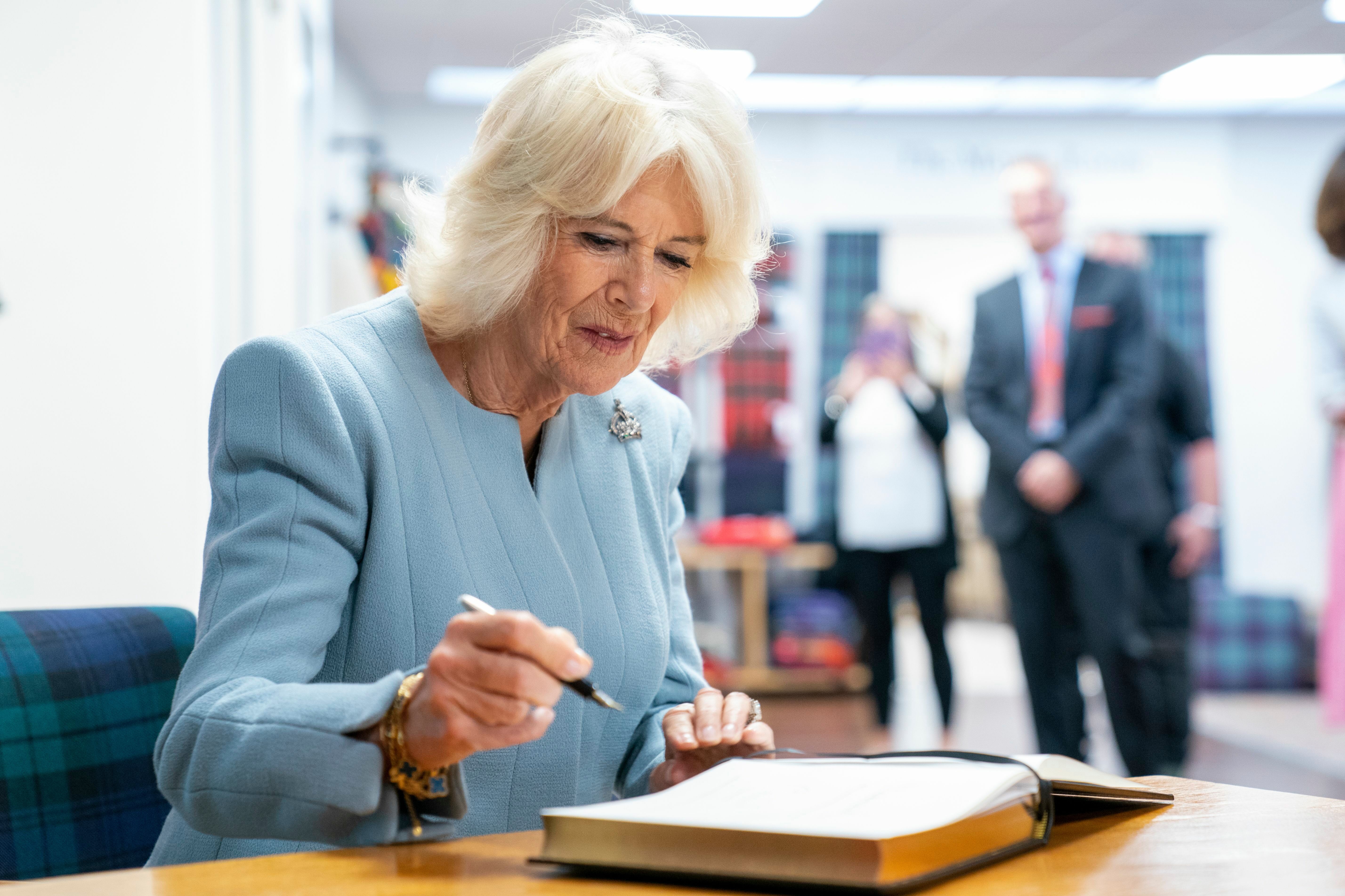 Queen Camilla sat down writing in a book, wearing her crown brooch pinned to a light blue dress