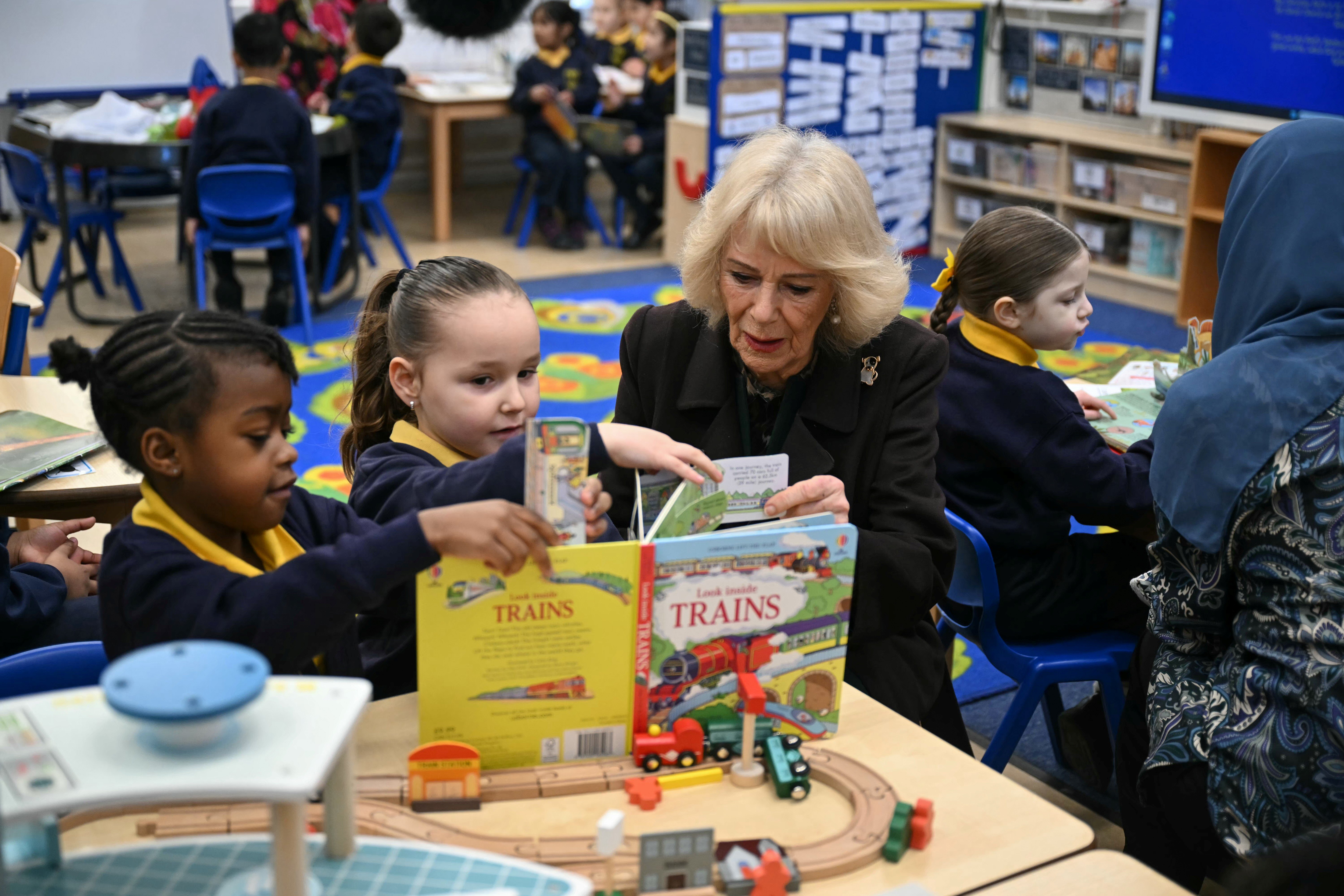 Queen Camilla sat down reading a book to two primary school children in uniform, Camilla is wearing a brown coat dress and a Jack Russel dog brooch