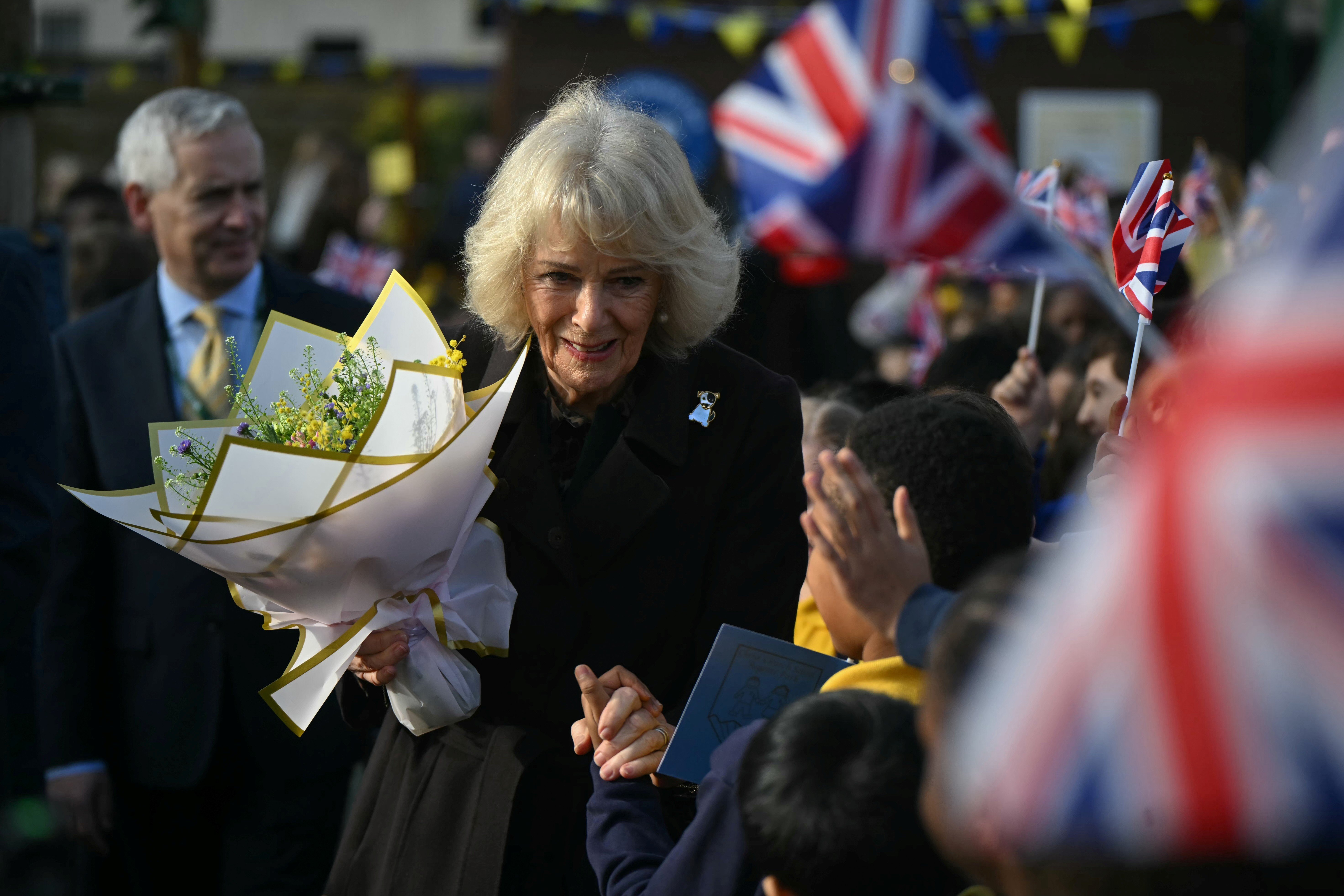 Queen Camilla collecting flowers from a crowd, wearing a brown jacket and her dog brooch