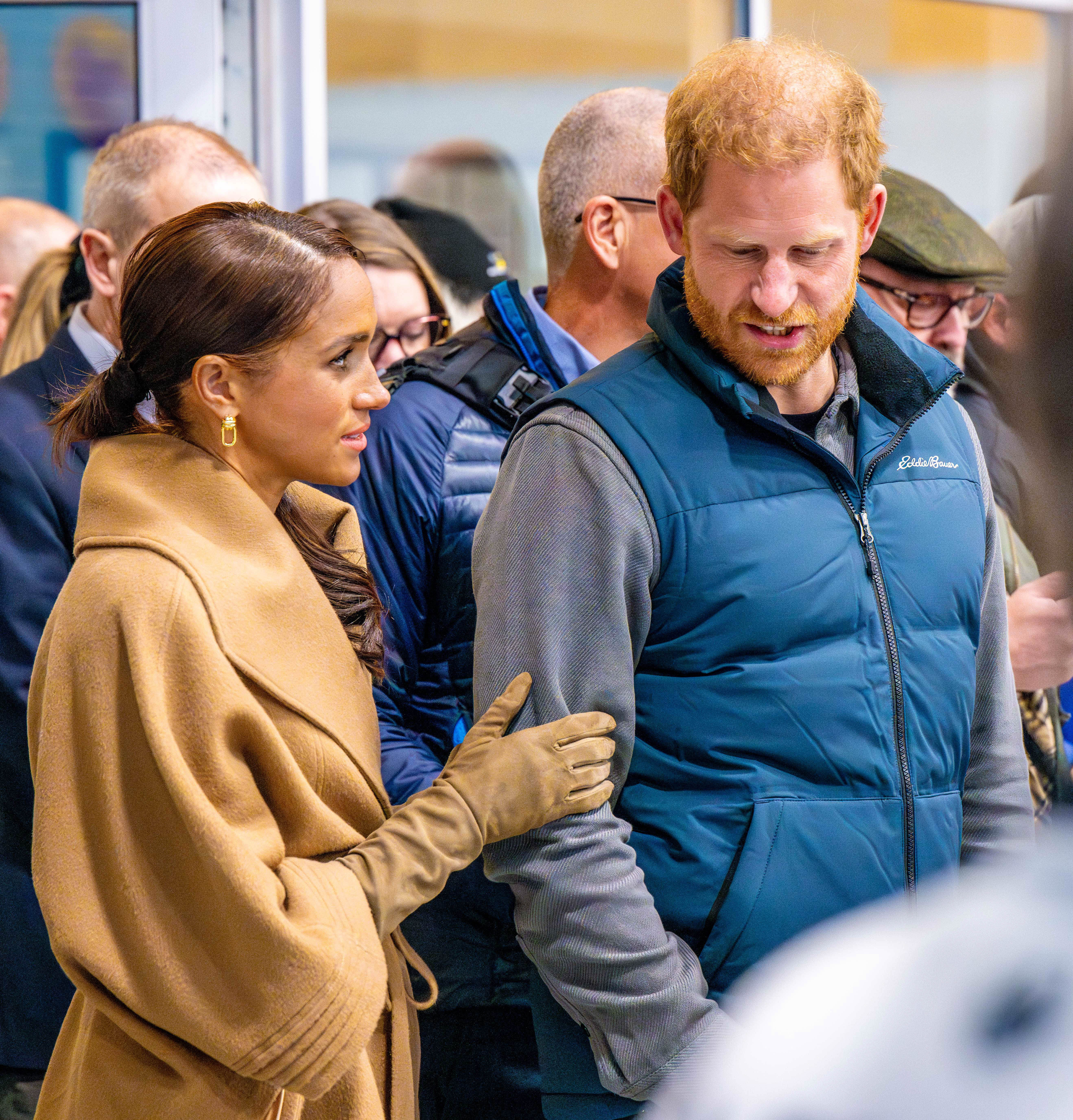 Meghan Markle touching Prince Harry's arms as he speaks with a disgruntled expression