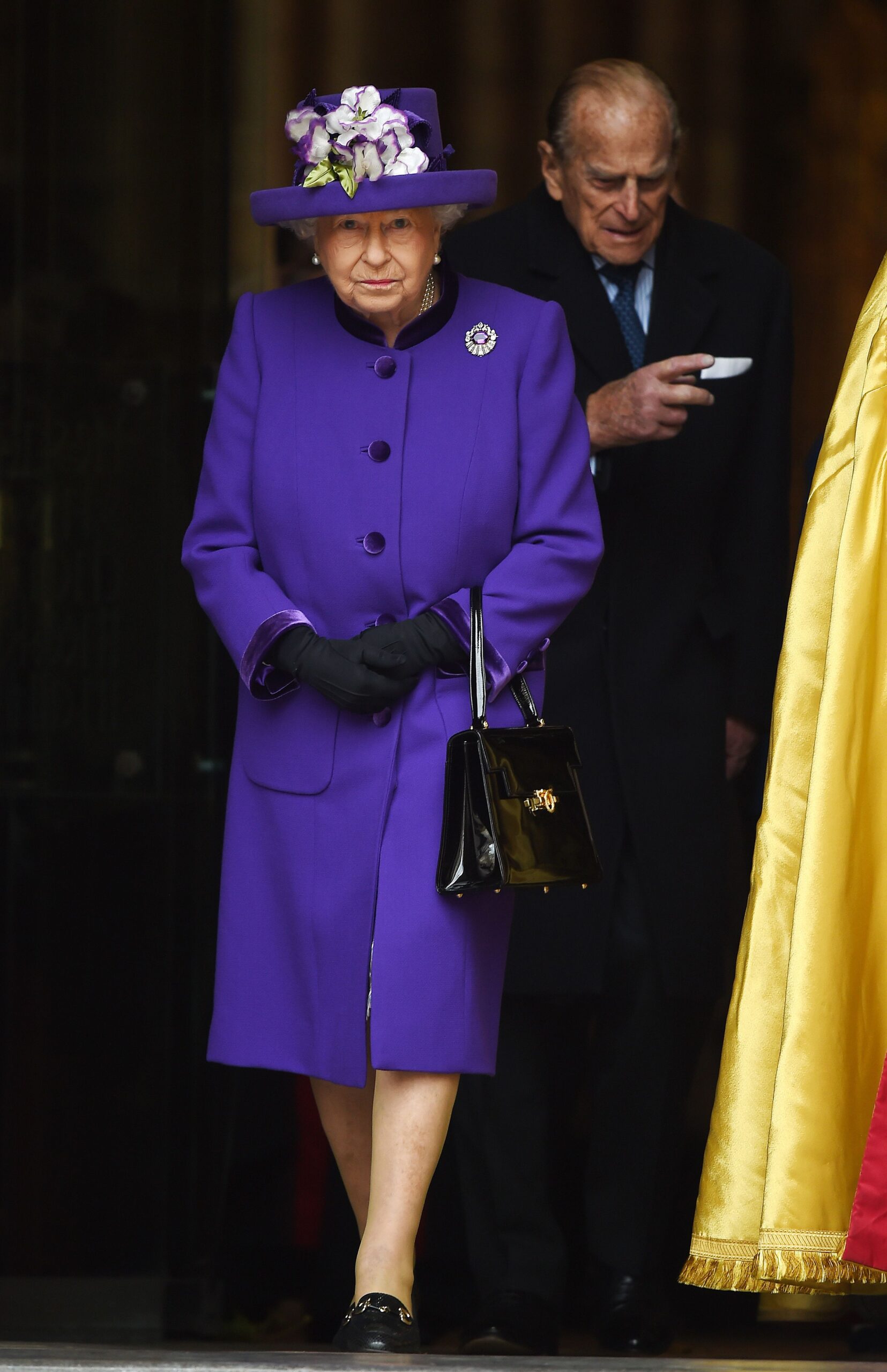 Queen Elizabeth II in purple outfit and Prince Philip behind her