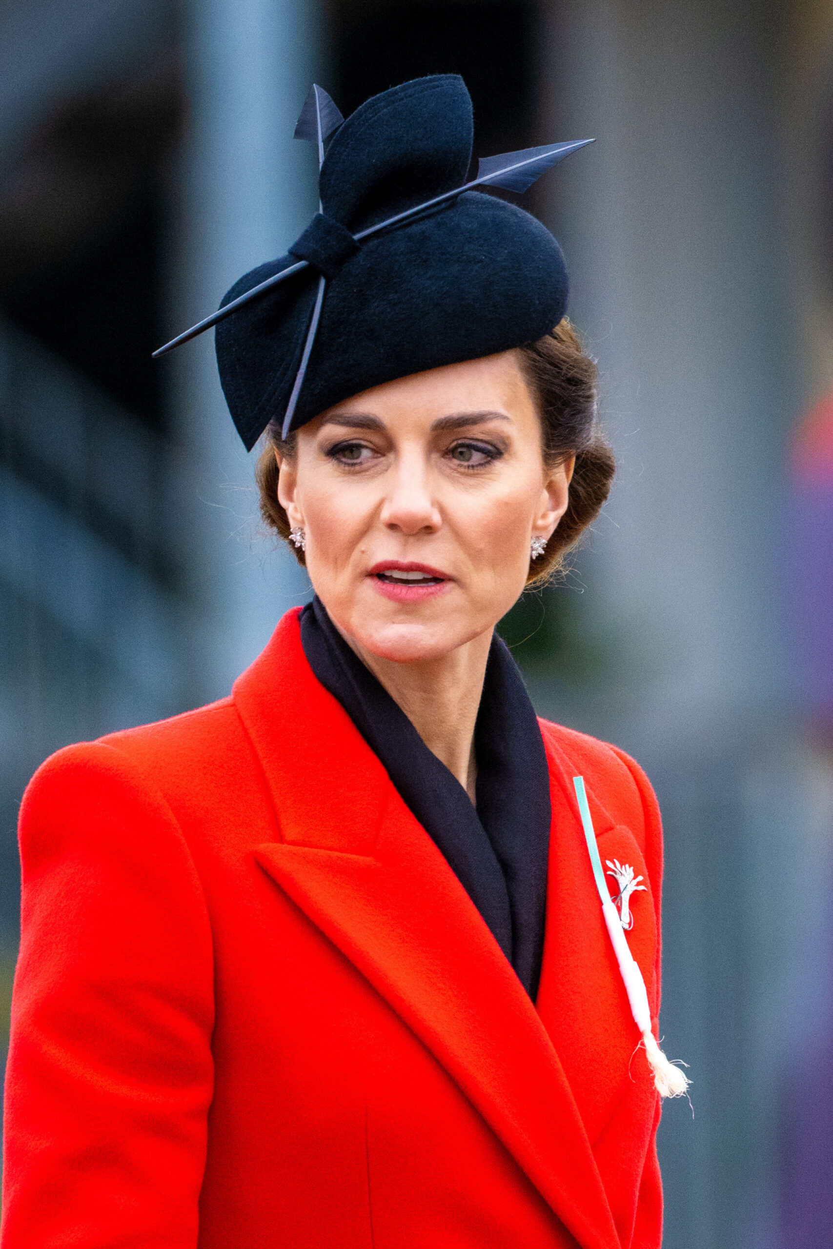 Princess Kate Middleton with a serious expression wearing a black hat and a red coat