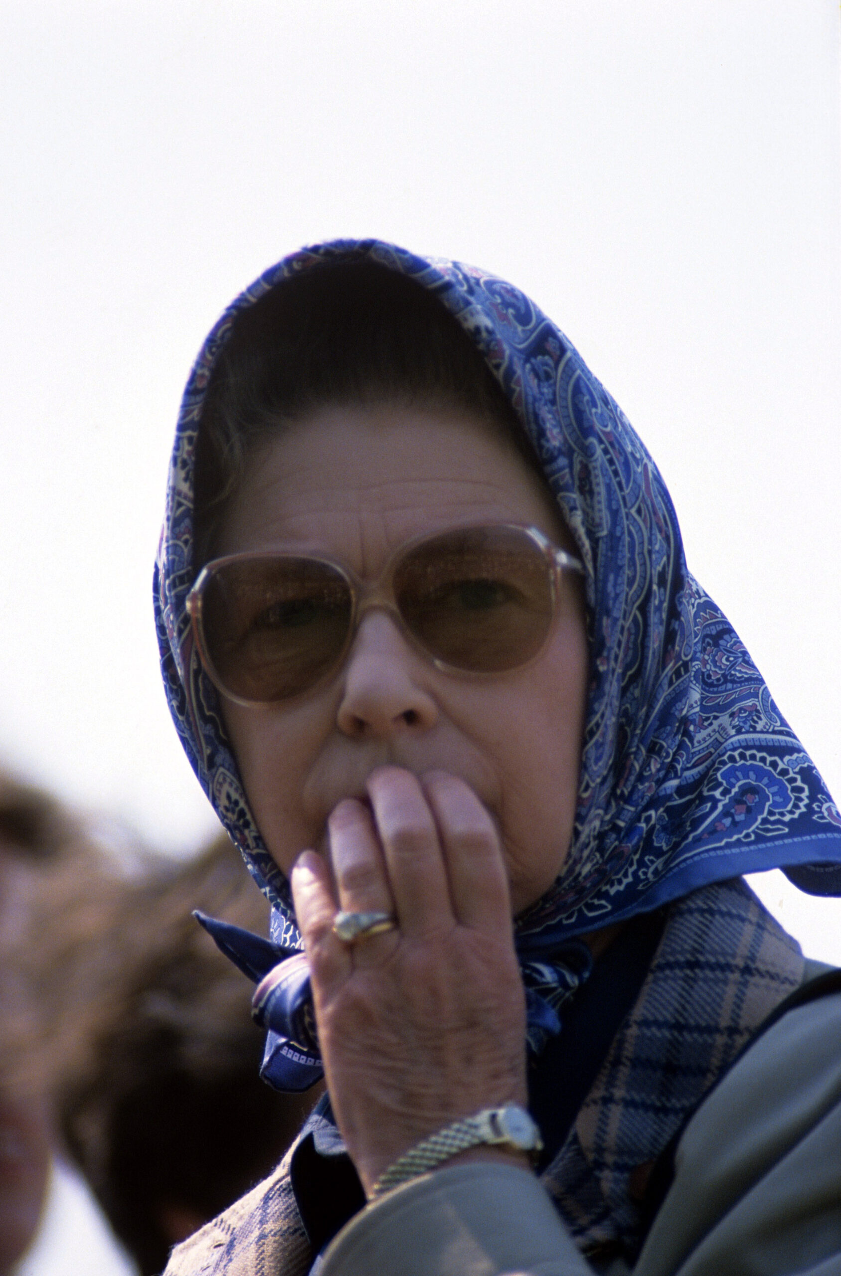 Queen Elizabeth wear sunglasses and a headscarf, touching her lips worriedly