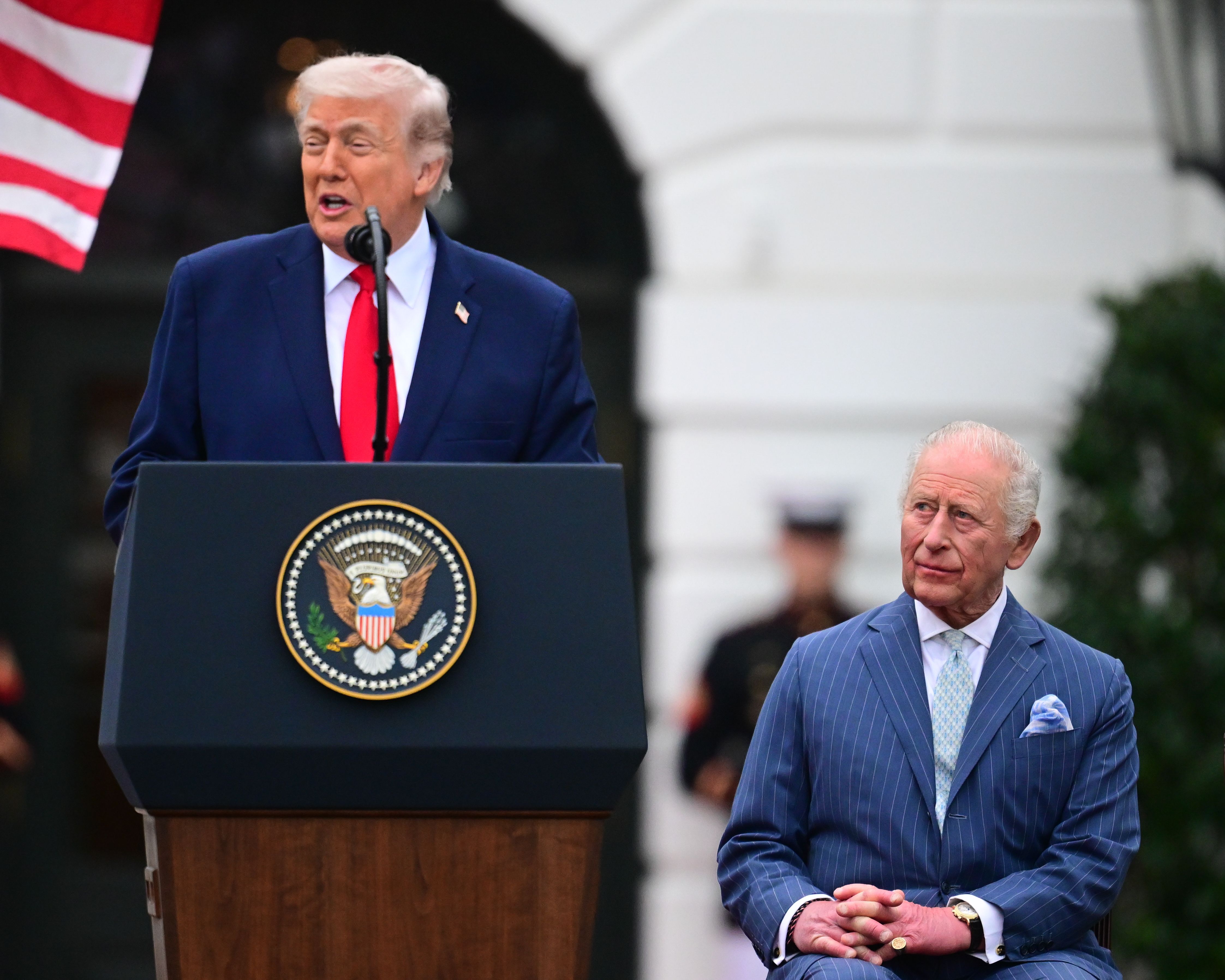 King Charles seated, frowning, as Donald Trump speaks at a podium